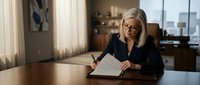 Professional blonde businesswoman in dark suit reviewing documents at executive desk in modern corporate office with city skyline view through windows.