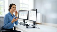 Professional Hispanic businesswoman eating healthy snack while working at dual monitor computer setup in modern bright office environment.