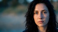 Young brunette woman with curly hair and freckles looking directly at camera with serious expression during golden hour lighting for portrait photography.