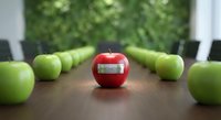 Red apple with organic label stands out among green apples on wooden conference table in modern boardroom with blurred greenery background for business concepts.