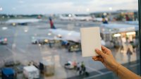 Person holding blank white card at airport terminal window with planes and ground vehicles visible below.