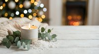 Cozy winter home decor with burning candle and eucalyptus on white wooden table. Warm Christmas lights and fireplace create festive holiday atmosphere.