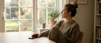 Young woman in cozy sweater sitting at wooden table by window, enjoying peaceful morning moment with coffee cup in bright, warm home interior.