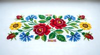 Traditional cross-stitch embroidery pattern with colorful floral motif featuring red roses, blue cornflowers and yellow sunflowers on white fabric.