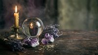 Mystical fortune telling setup with crystal ball, burning candle, amethyst crystals and dried lavender on wooden table for spiritual divination practices.