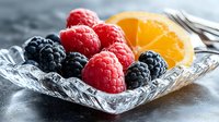 Fresh ripe raspberries and blackberries with orange slice served in elegant crystal glass bowl on dark background, macro food photography.