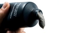 Caucasian male hand holding black cosmetic scrub bottle with product pouring out onto white background for skincare and beauty product marketing.