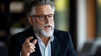 Confident mature businessman with gray beard and glasses gesturing while speaking during professional meeting, wearing classic suit and white shirt.