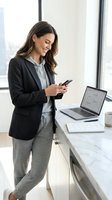 Young professional woman of mixed ethnicity wearing business casual attire, holding smartphone while working at modern office desk with laptop and documents, suitable for corporate and business themes.