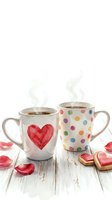 Two ceramic mugs with hot beverages on white wooden surface, one featuring red watercolor heart, other with colorful polka dots, surrounded by rose petals and heart-shaped cookies.