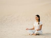 Young woman of South Asian descent in white casual clothing practicing meditation and mindfulness on sandy beach during daytime for wellness and relaxation content.