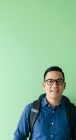 Young Asian male wearing eyeglasses and blue denim shirt with black backpack, smiling against mint green background for portrait and professional headshot use.