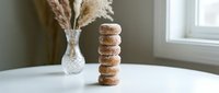Stack of six glazed donuts balanced on white table near window with pampas grass vase creating minimalist food photography composition for bakery marketing.