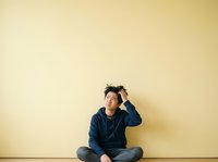 Young Asian male, casual appearance, wearing dark blue sweatshirt and jeans, sitting cross-legged against plain yellow wall, hand to head in thoughtful or confused pose, studio setting.