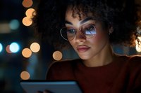 Young woman of mixed ethnicity wearing round eyeglasses and burgundy sweater, focused on tablet device at night with bokeh city lights in background. Evening work or study scene.