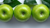 Fresh green apples with water drops and leaves arranged in row on light gray background. Healthy organic fruit macro photography for food design.