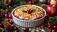 Festive cranberry crumble dessert in white ceramic baking dish surrounded by fresh cranberries, pine cones, apples and evergreen branches on rustic wooden surface.