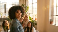Happy African American woman with curly hair smiling in bright office space with diverse female colleagues in background during team meeting or workshop.