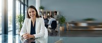 Professional businesswoman in white blazer smiling confidently at modern office desk with colleagues working in background corporate workplace.