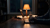 Man working late at night on laptop computer at wooden desk with warm table lamp creating cozy home office atmosphere for productivity and focus.