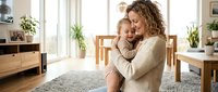Loving Caucasian mother with curly blonde hair embracing her smiling toddler son in cozy modern living room with natural light and plants.
