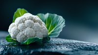 Fresh white cauliflower head with green leaves on dark wet surface against blurred background. Dramatic lighting highlights vegetable texture and natural form.