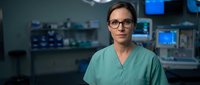Professional female surgeon in green scrubs and glasses standing confidently in modern operating room with medical equipment and monitors in background.