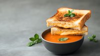 Grilled cheese sandwich stacked on black bowl with creamy tomato soup garnished with fresh basil leaves on gray background, comfort food styling.