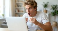 Young Caucasian man with blonde hair showing shocked surprised expression while looking at laptop screen in modern home office interior.