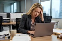 Stressed middle-aged Caucasian businesswoman holding head while working on laptop in modern office, experiencing work pressure and fatigue during busy day.