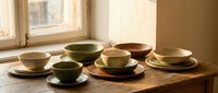Handmade ceramic bowls and plates in earthy tones displayed on wooden table near window. Artisan pottery collection for kitchen dining decor.