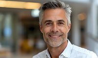 Confident mature Caucasian businessman with gray hair and stylish stubble wearing white shirt smiles warmly at camera in modern office environment. Professional corporate portrait.