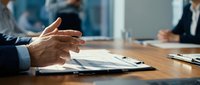 Businessman gesturing during corporate meeting at wooden table with documents and colleagues in background, suitable for business communication and teamwork concepts.