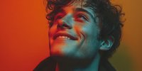 Young man with curly hair smiling under dramatic red and blue lighting, creating a vibrant mood portrait suitable for music promotion or youth culture content.