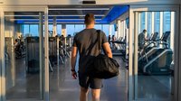 Athletic man entering modern fitness center with gym bag, ready for workout session in bright contemporary health club with cardio equipment.