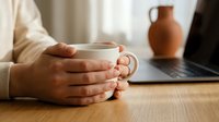 Hands holding white ceramic mug at wooden desk with laptop and clay pitcher creating cozy home office workspace for remote work and morning coffee rituals.