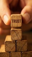 Hands carefully placing wooden cube with TRUST text on pyramid of blocks symbolizing business reliability, teamwork and foundation building concepts.
