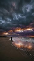 Solitary surfer in wetsuit standing on sandy beach at dramatic sunset with stormy clouds and golden light reflecting on ocean waves, suitable for travel and lifestyle content.