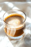 Fresh coffee with creamy foam in clear glass on white textile background, natural morning light creates soft shadows. Aromatic beverage for cafe menu photography.