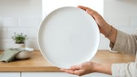 Empty white plate held by female hands with brown skin tone in modern kitchen setting, suitable for food styling and culinary content.