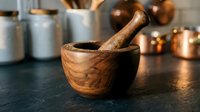 Wooden mortar and pestle on dark kitchen counter with copper cookware and storage jars in background for cooking and food preparation.