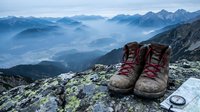 Worn hiking boots on rocky mountain summit overlooking misty alpine valley landscape at dawn with layered peaks and morning fog below.