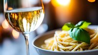 Creamy pasta dish with fresh basil garnish served alongside chilled white wine glass, creating warm ambiance with soft bokeh lights in background.