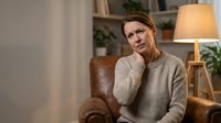 Middle-aged Caucasian woman experiencing neck pain sitting in leather armchair at home, touching sore area with concerned expression in cozy living room.