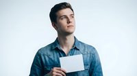 Young man with dark hair and light skin tone wearing a blue denim shirt, holding a blank white card or paper against white background for mockup and branding purposes.
