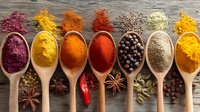 Colorful spices and herbs in wooden spoons arranged in row on rustic wooden background, including turmeric, paprika, chili, star anise, and peppercorns.