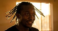 Young African American man with dreadlocks smiling warmly in golden sunlight streaming through window, creating atmospheric portrait with natural lighting.