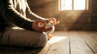 Person in warm clothing holding wooden rosary beads while sitting on wooden floor in sunlit room, conveying spirituality, faith, prayer, and peaceful meditation practice.