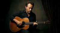 Middle-aged Caucasian man playing acoustic guitar in dimly lit room with dramatic lighting and curtains for music and entertainment content.