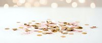 Scattered golden and rose gold confetti pieces on white surface with soft bokeh lights creating festive celebration atmosphere for party decorations.